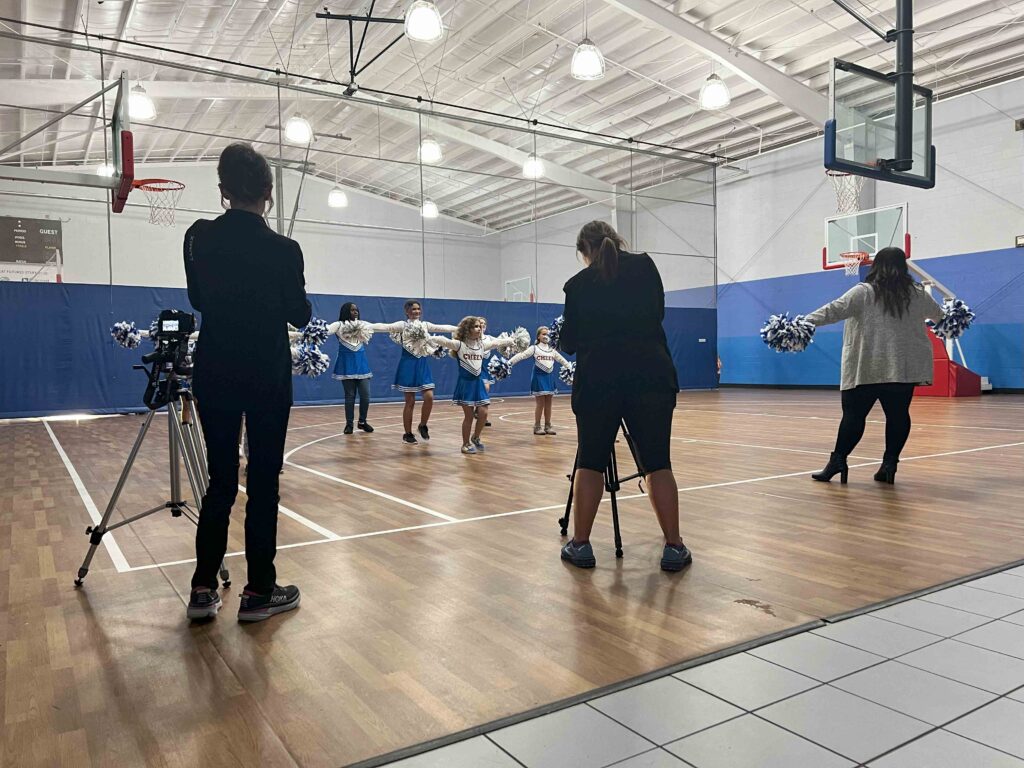 Behind the scenes of a video production of a fundraising video for a young girl's charity filming the girls doing a cheer routine on a basketball court.