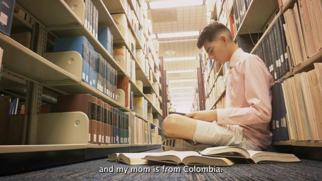 A young college student sitting on the floor amongst the bookshelves in the library with books open all around him.
